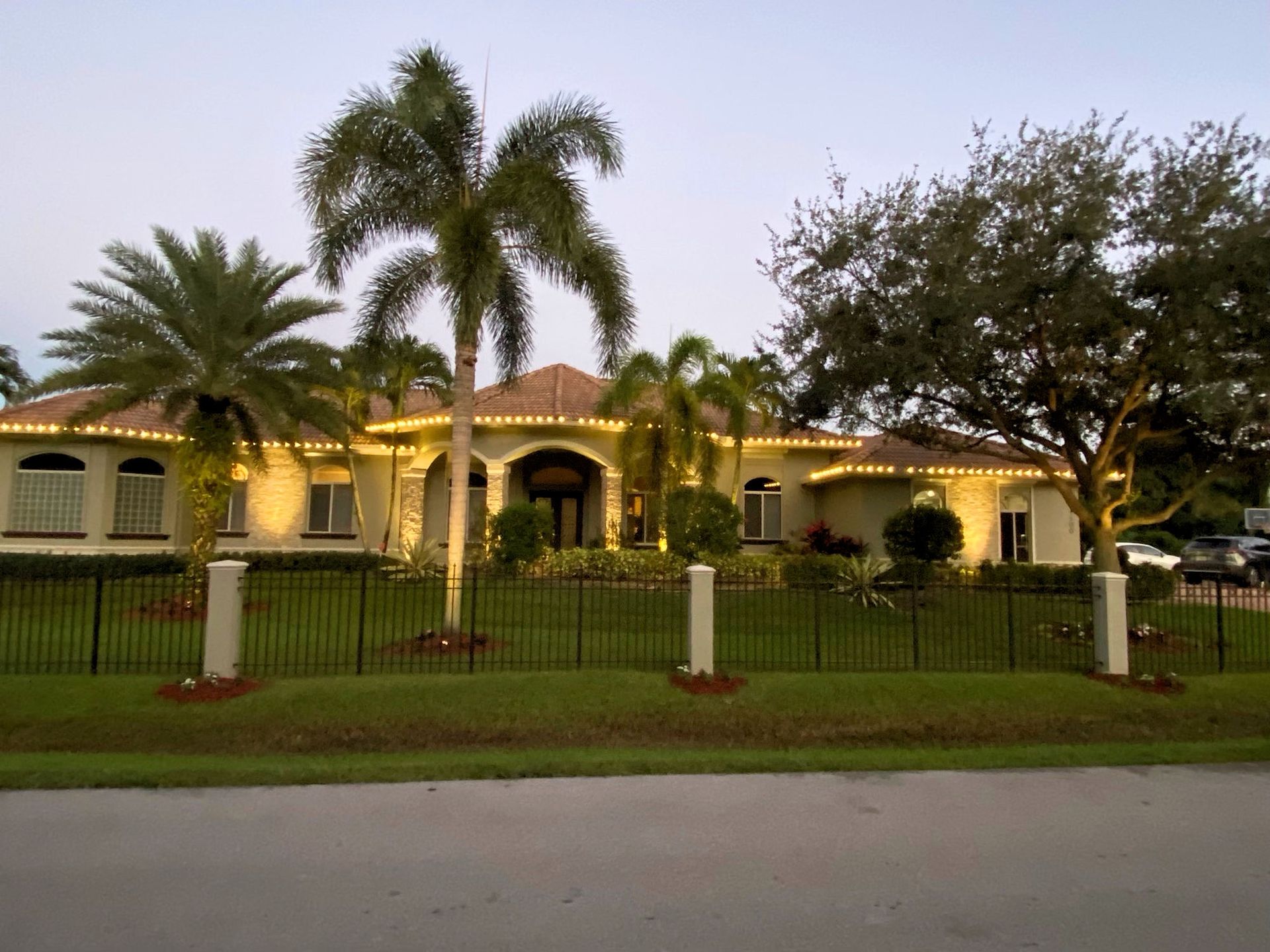 A single-story, tan-colored house at twilight, illuminated by decorative roofline lights, with palm trees and a lawn.