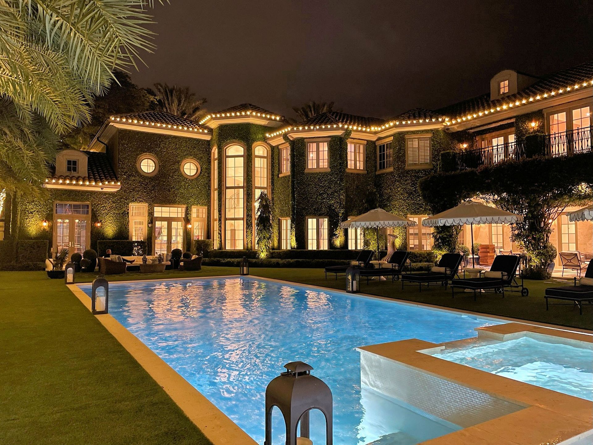 A lit mansion at night overlooks a backyard swimming pool and hot tub with green landscaping and patio umbrellas.