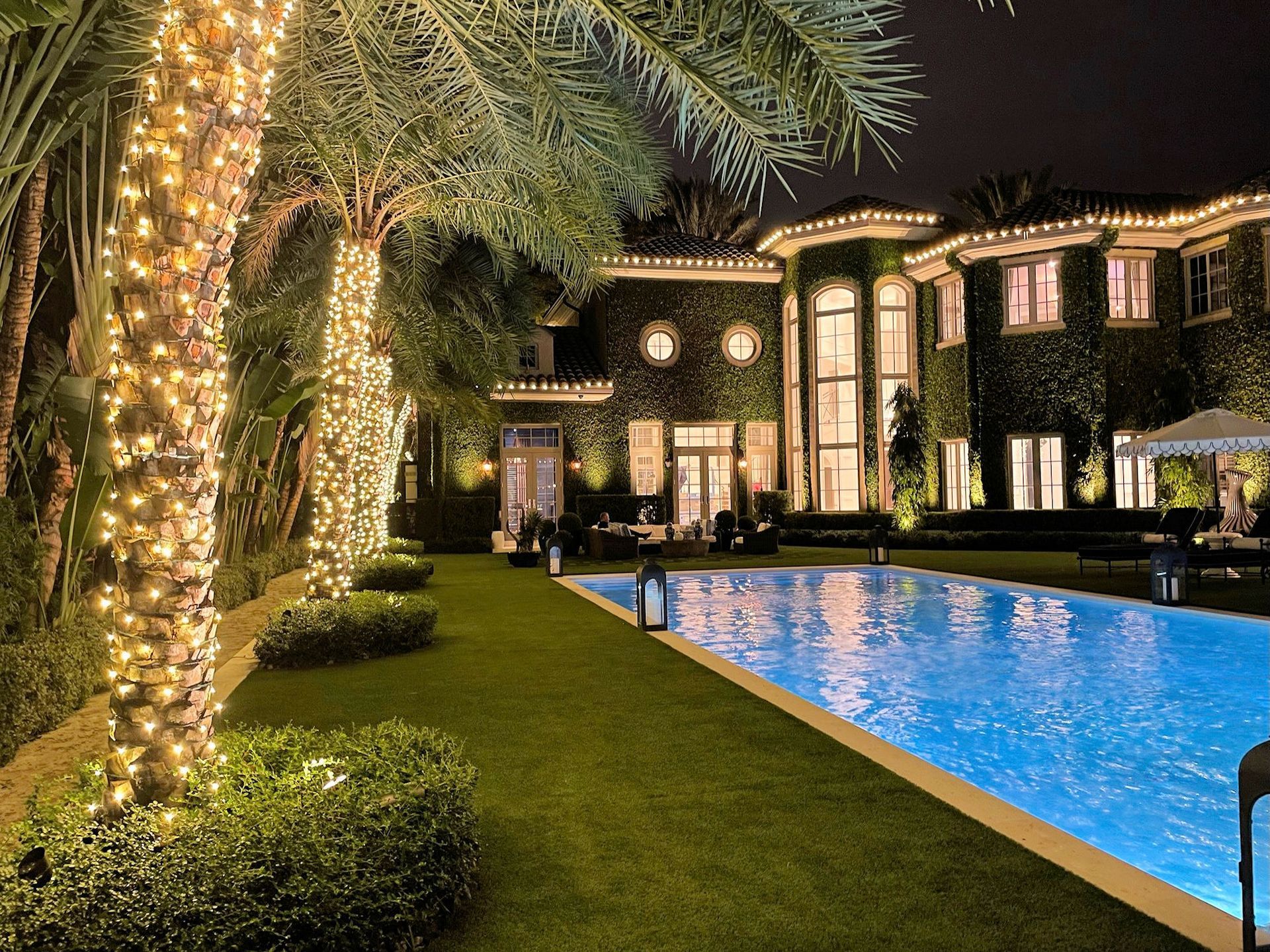 A luxurious mansion with ivy-covered walls at night, illuminated by string lights on palm trees beside a glowing pool.