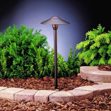 A bronze-colored, mushroom-style path light stands in a garden bed with wood mulch, green shrubs, and a stone border.