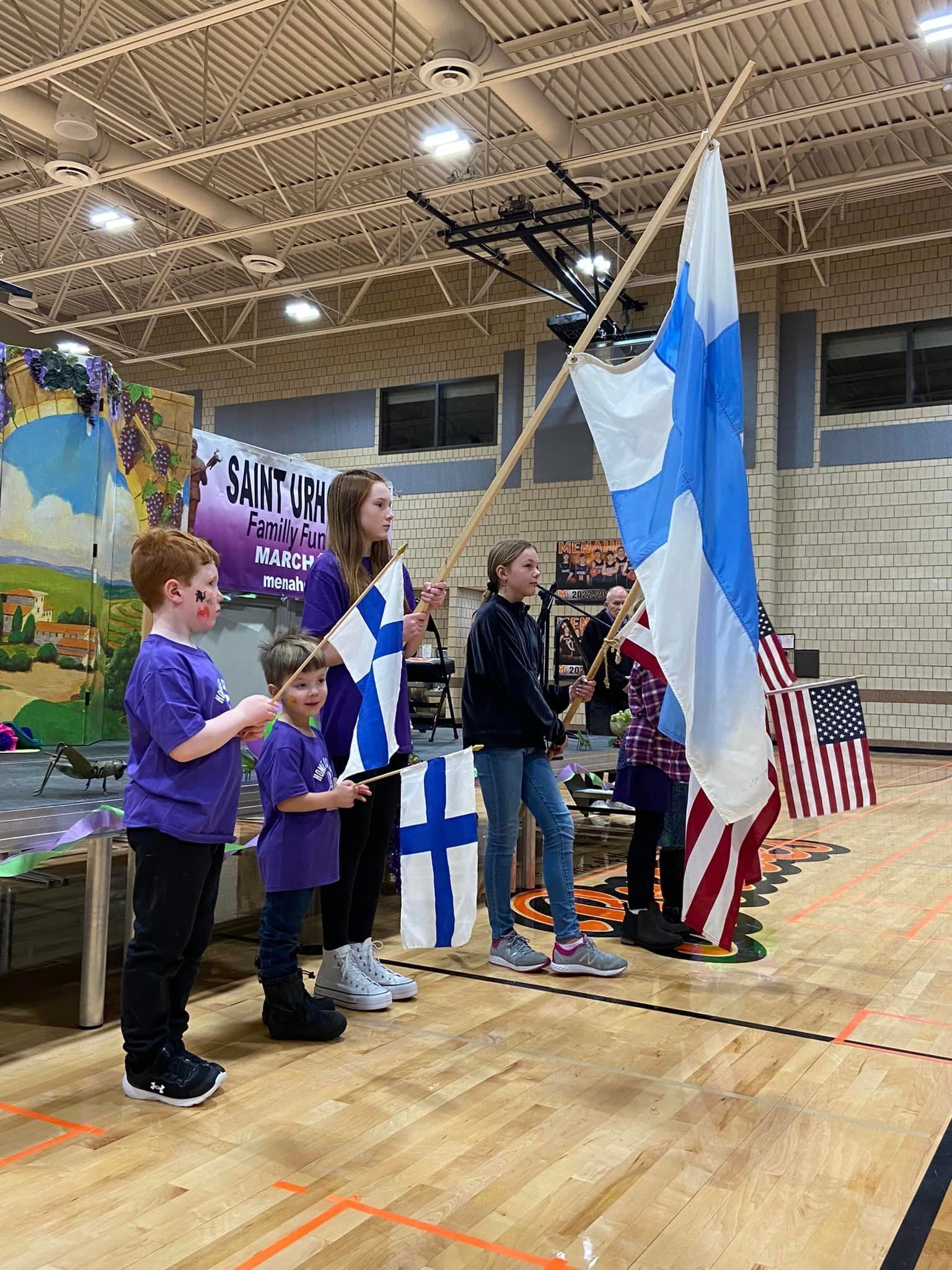 Children hold Finnish flags and stand near a US flag in a gymnasium for St. Urho Days.