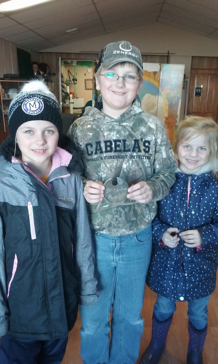 Three children stand together in an indoor space; one holds a small, dark object while they all smile at the camera.