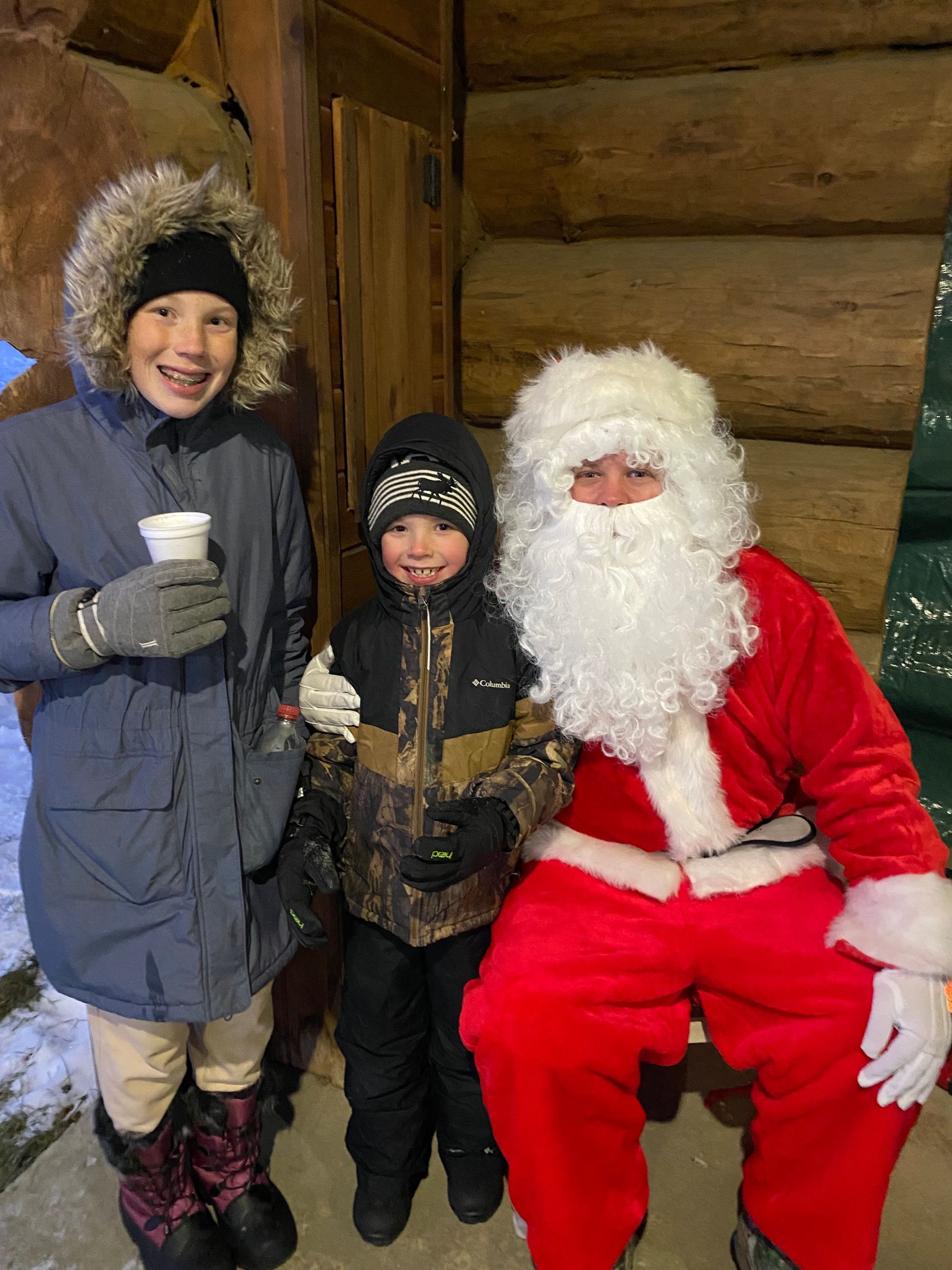 Two children in winter coats stand next to Santa Claus in a red suit inside a rustic log building.