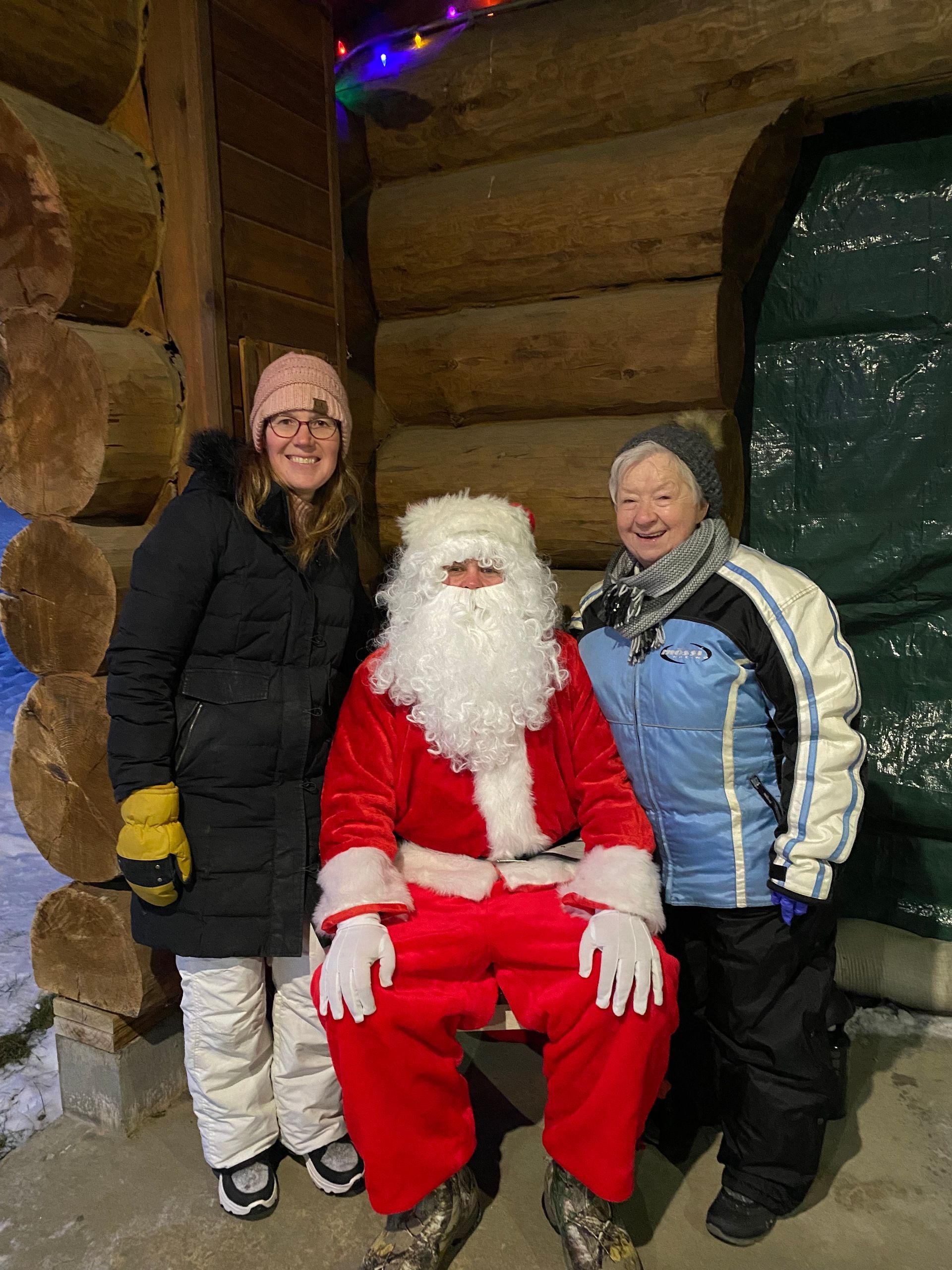 Two people stand on either side of Santa Claus in front of a rustic log cabin decorated with holiday lights.