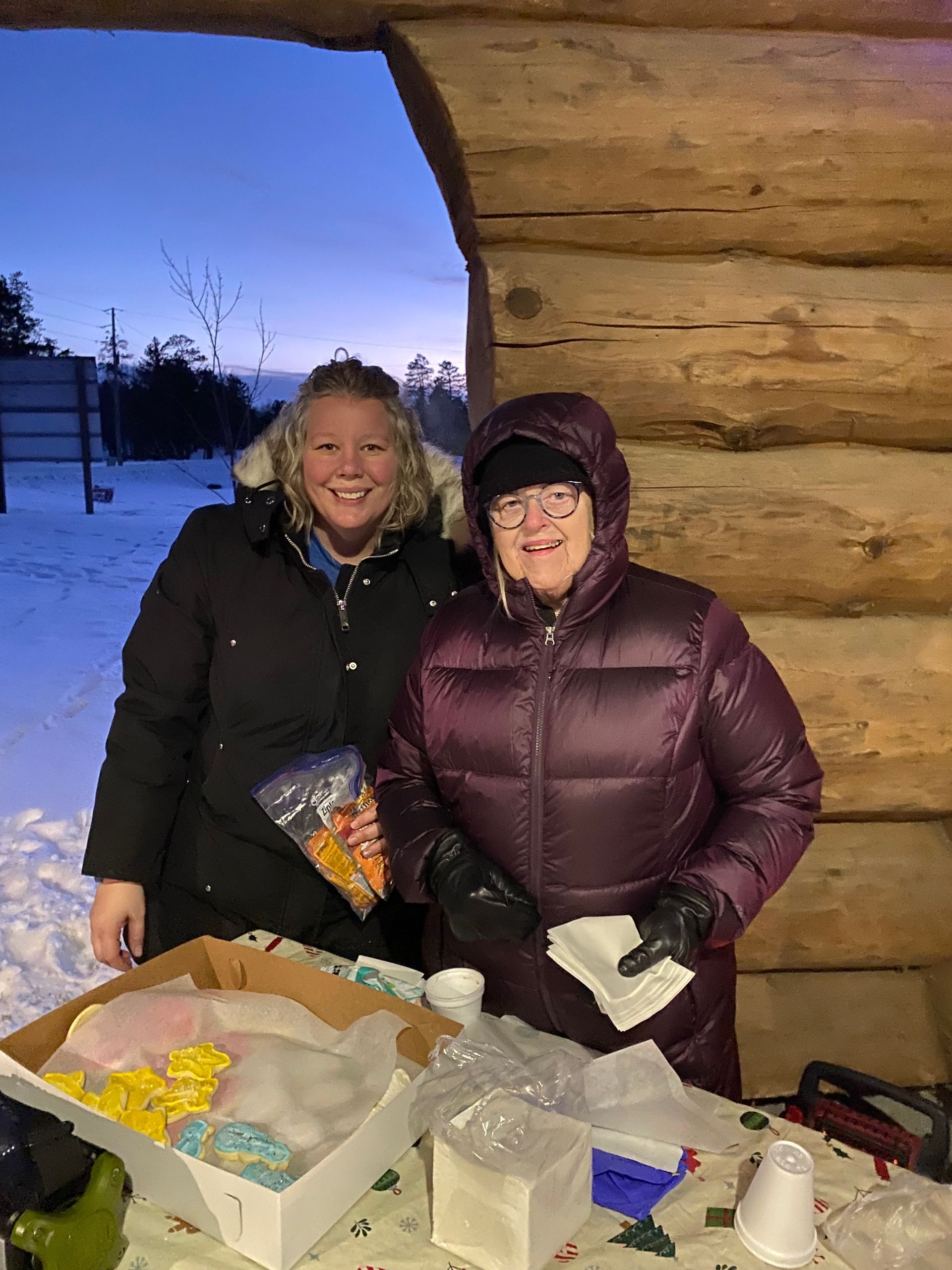 Two people smiling behind a table of treats in front of a log structure outdoors in the snow at dusk.