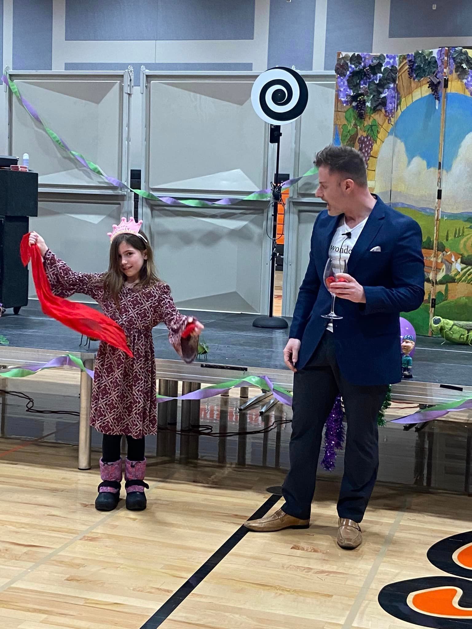 A performer in a blue blazer watches a child in a floral dress holding a red scarf on a stage with colorful decorations.