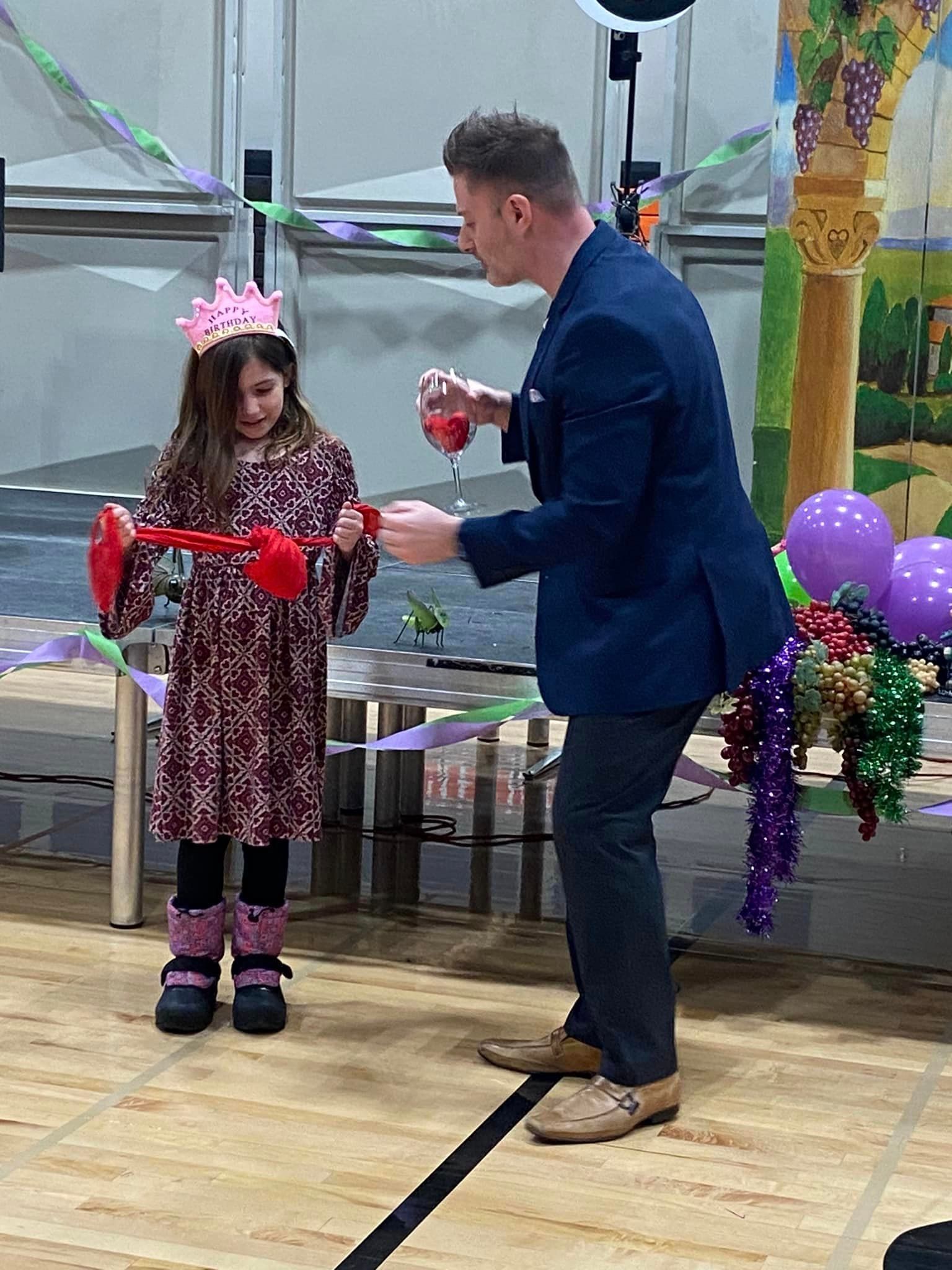A magician in a blue suit performs a trick with red ribbons for a child wearing a crown at a decorated event.