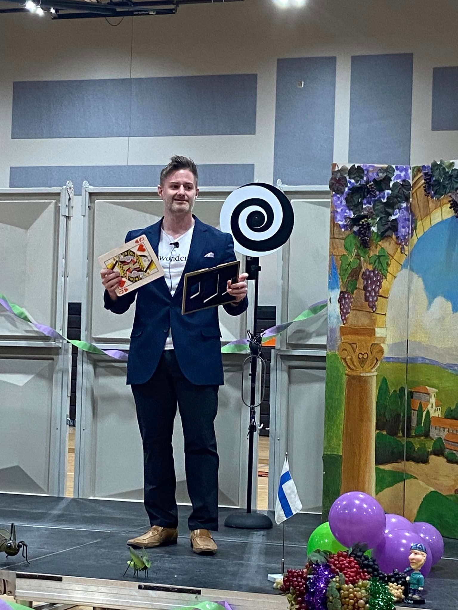 A man in a navy suit stands on a stage holding a small sign and a plaque, with a spiral backdrop and balloons nearby.
