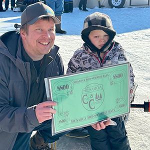 A smiling adult and a child in winter gear hold a $500 Menahga Fishing Derby prize certificate in the snow.