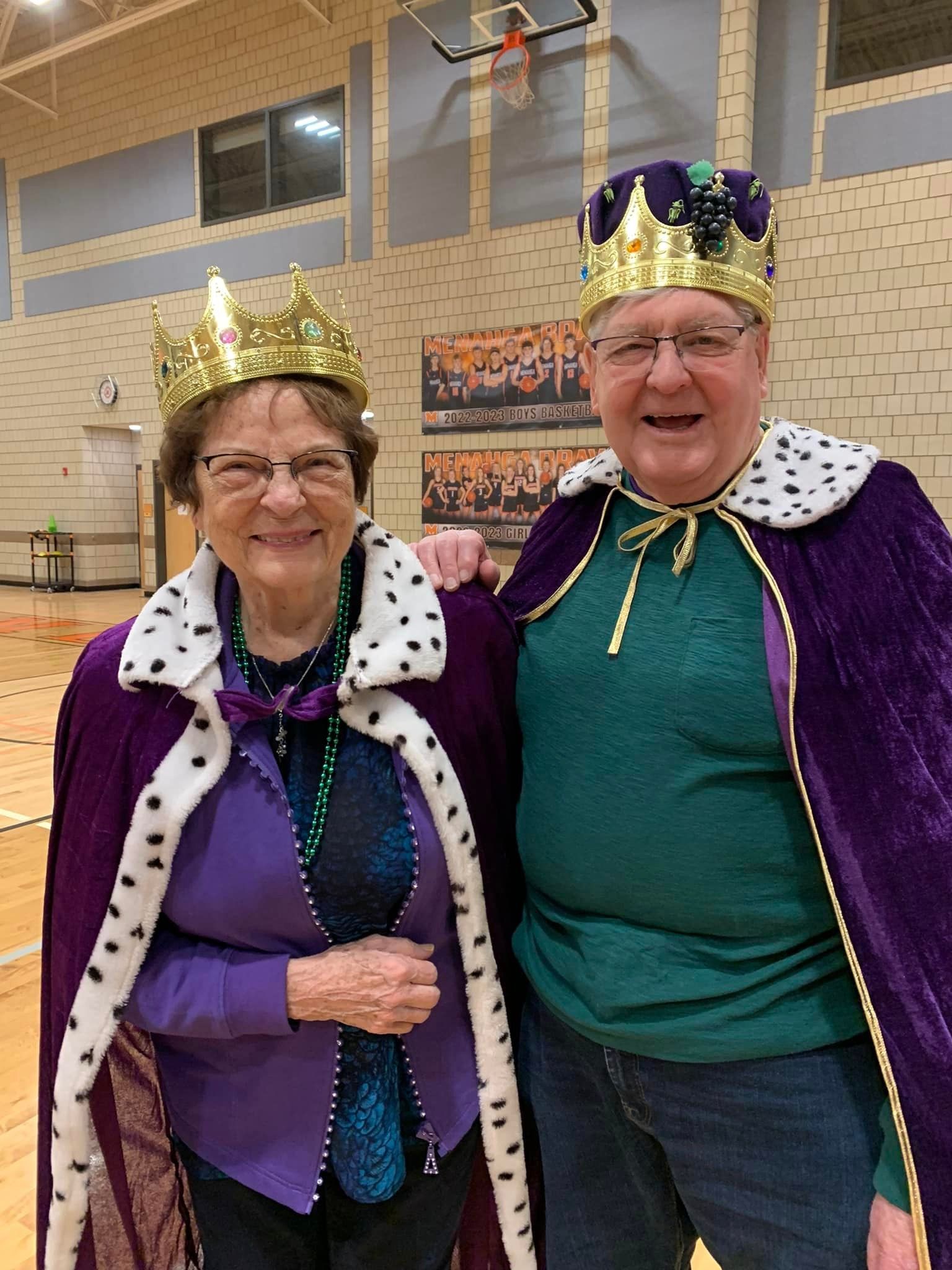 Two people wear gold crowns and matching purple capes with white fur trim, smiling in a gymnasium setting.