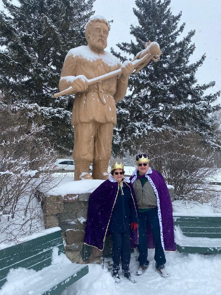 Two people wearing gold crowns and purple capes pose in a snowy park next to a tall wooden statue holding a wooden tool.