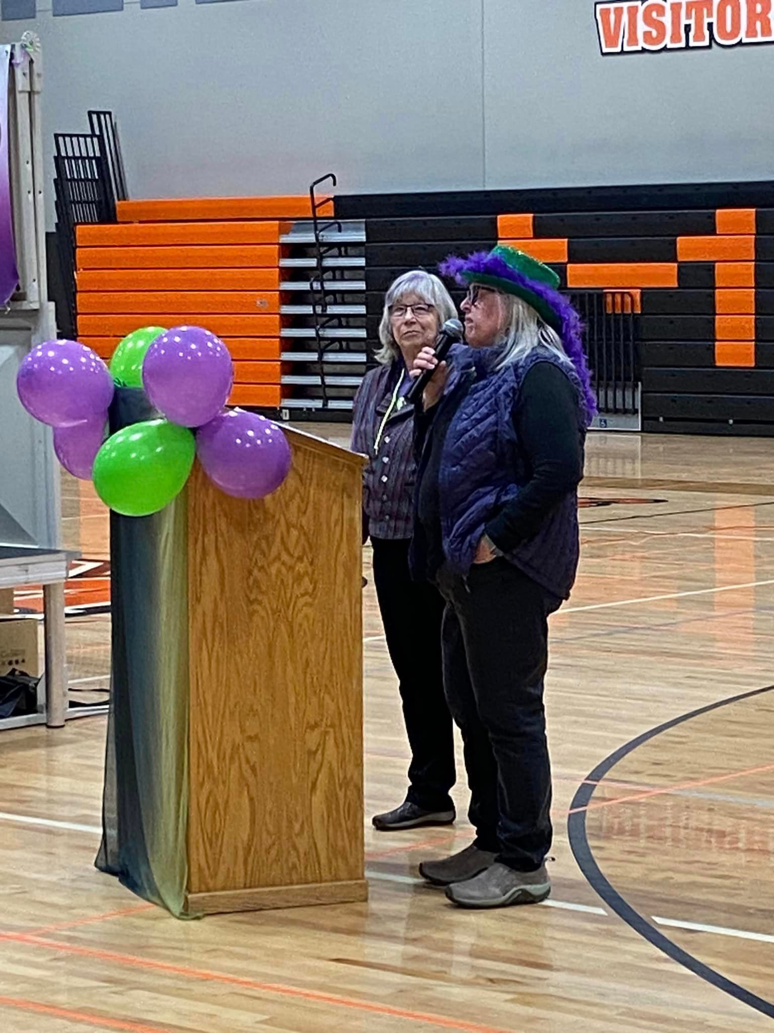 Two people stand behind a wooden podium at a school basketball court, decorated with green and purple balloons.