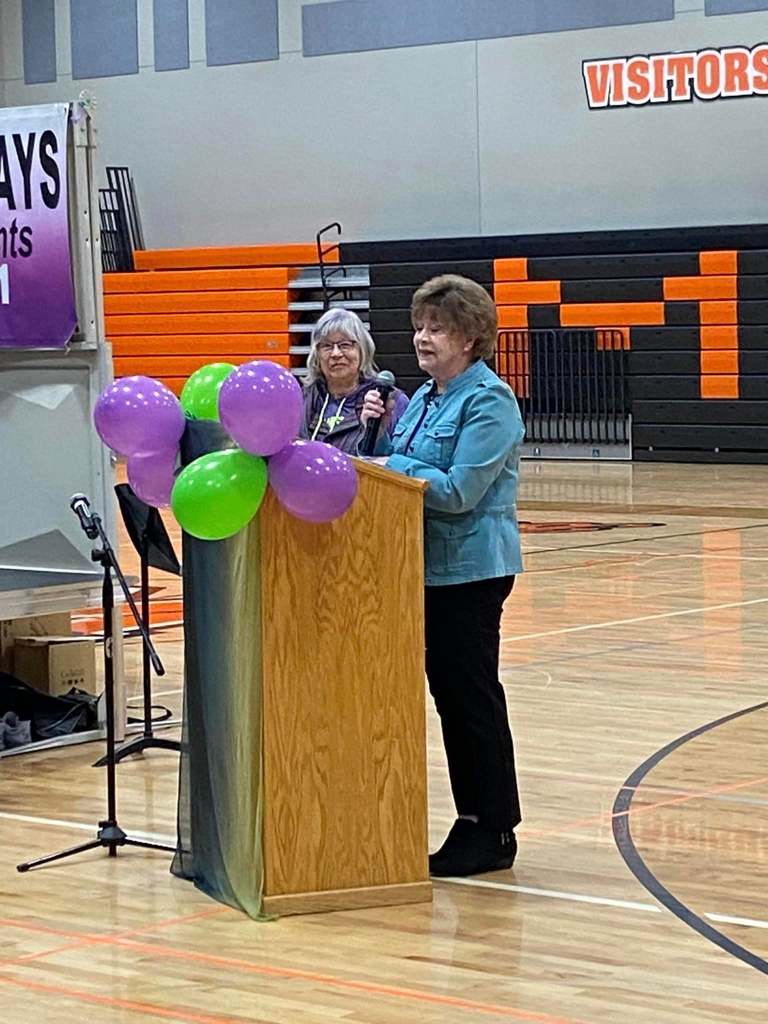 A person speaks at a wooden podium in a gymnasium, with another person behind them and balloons in front.