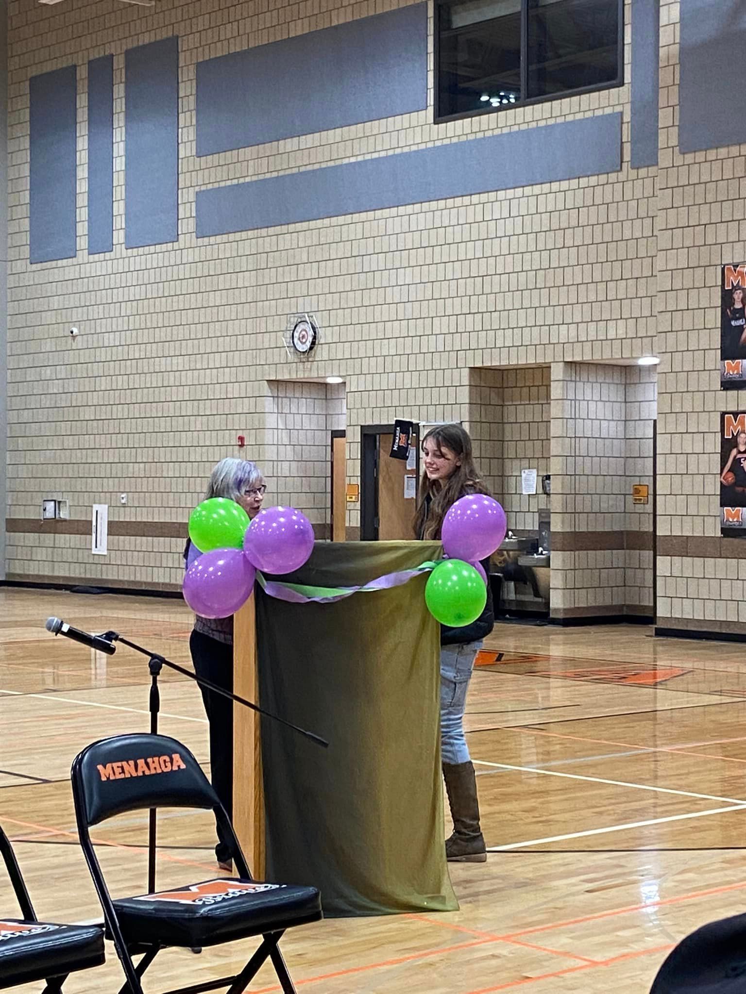 Two people stand behind a podium draped in green cloth, decorated with green and purple balloons, inside a gymnasium.