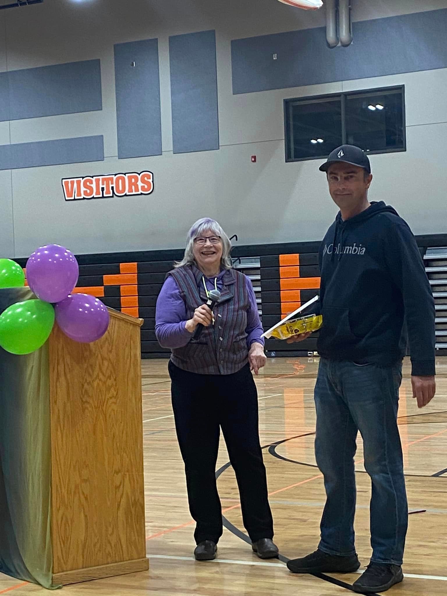 A person in a purple shirt stands at a wooden podium with balloons, while another person holds a trophy in a gym.