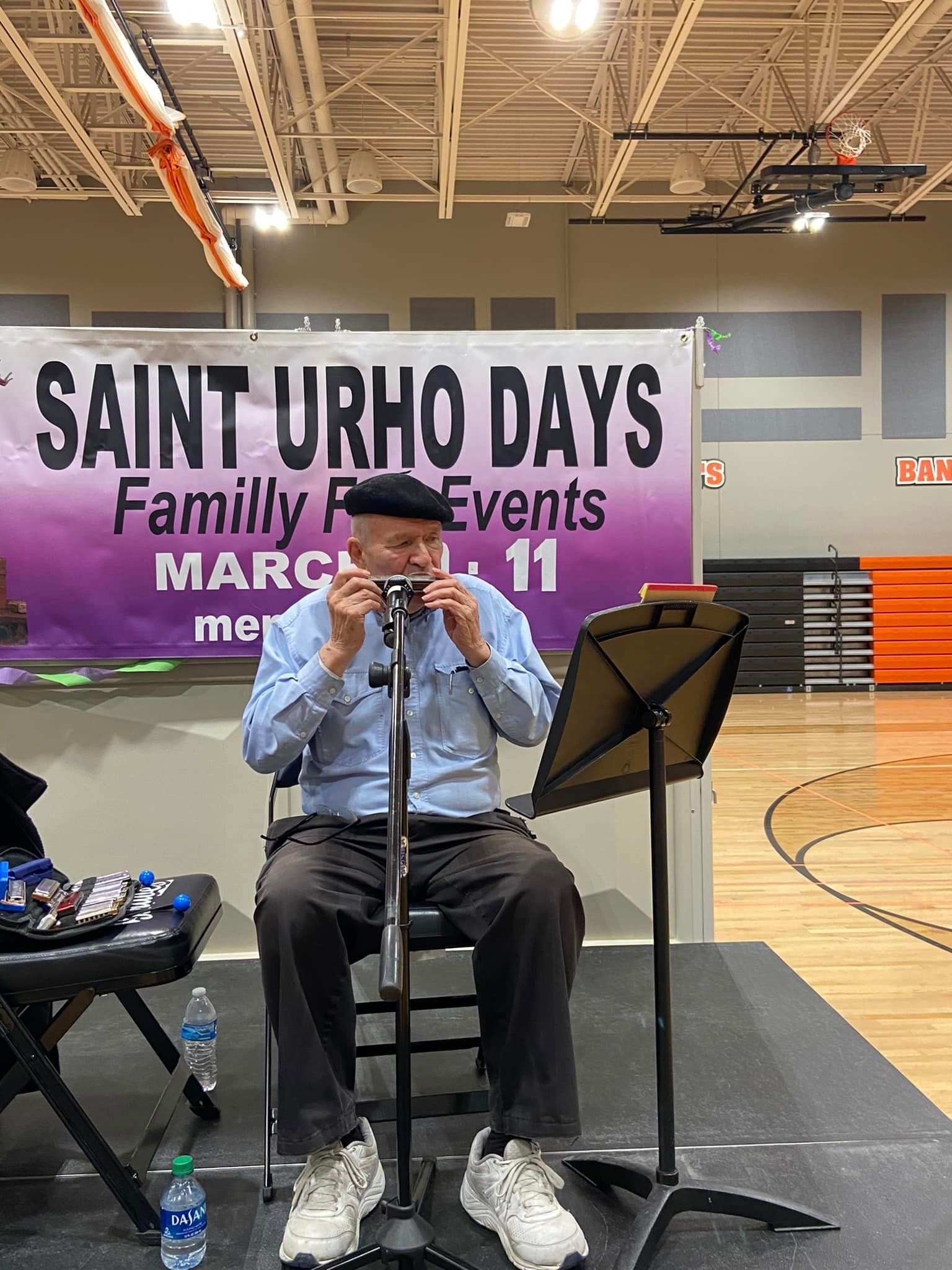 A person in a blue shirt and black beret plays a harmonica in front of a Saint Urho Days banner in a gymnasium.