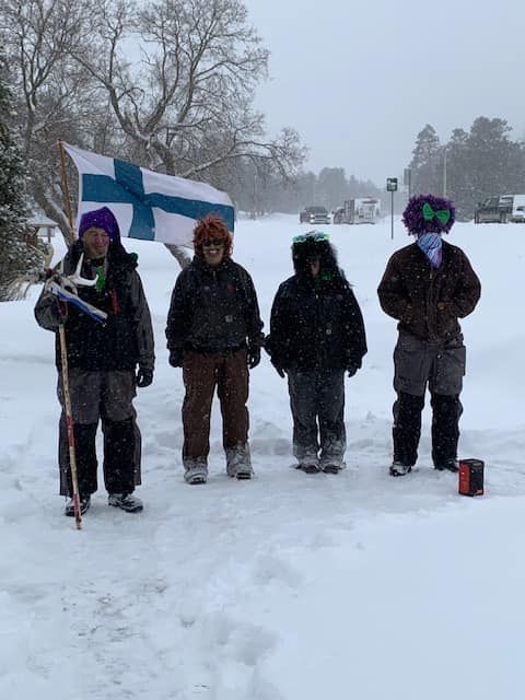 Four people wearing winter gear and colorful wigs stand in the snow, one holding a Finnish flag on a staff.