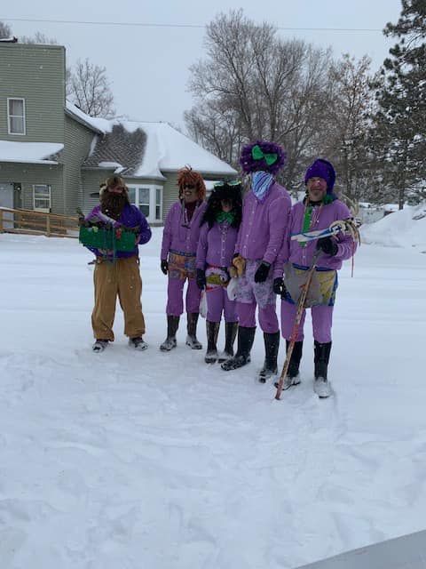 Five people in coordinated purple outfits and fun hats stand together in a snow-covered yard in front of a house.