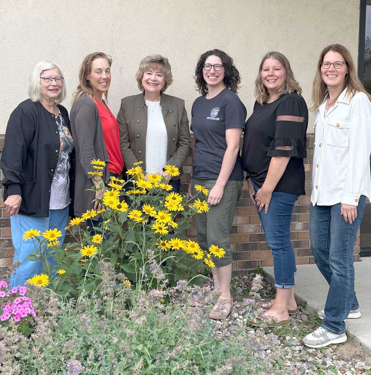 Six people stand in a line against a brick and stucco wall, posing behind a patch of yellow sunflowers and purple flowers.