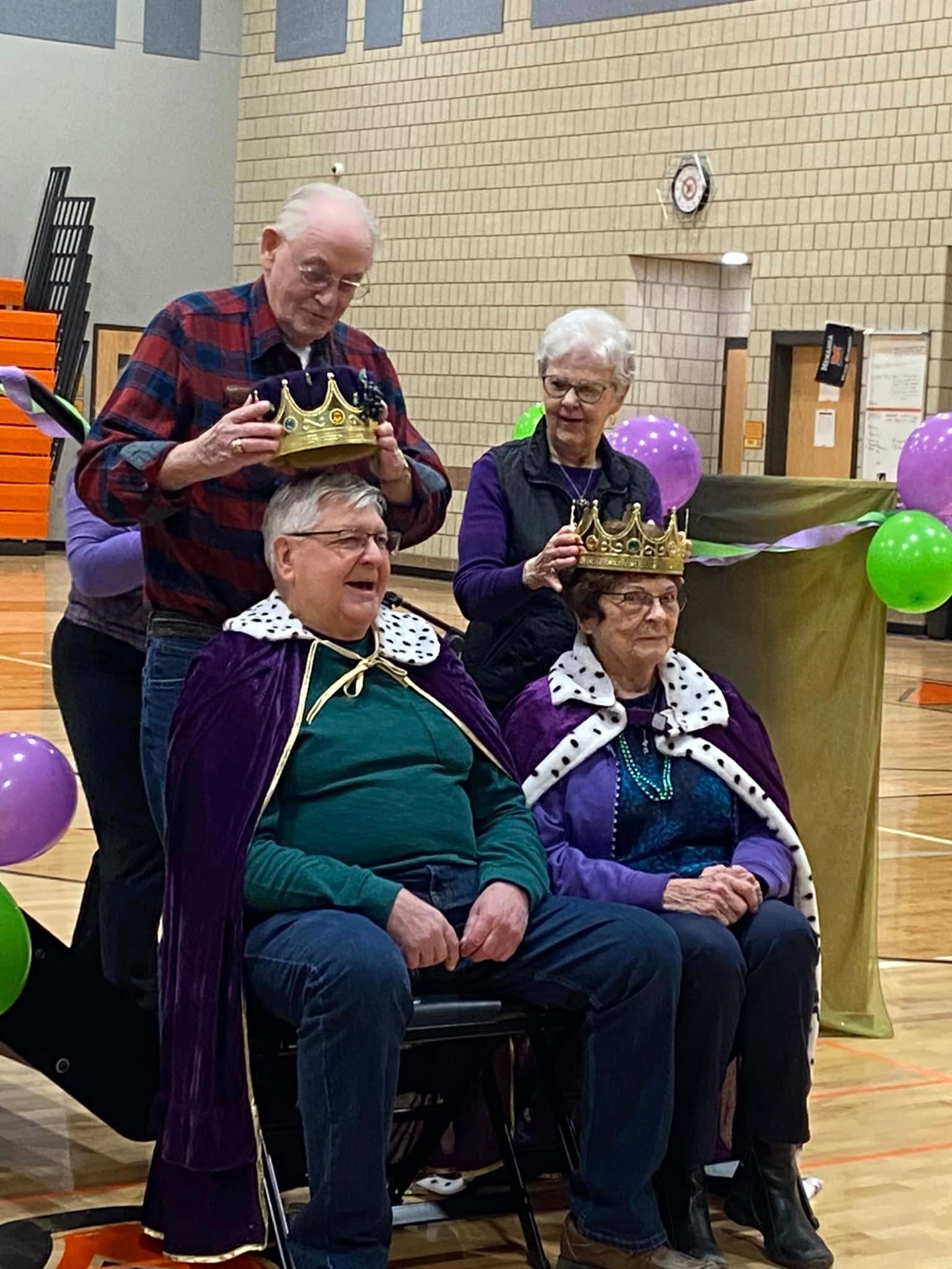 Two people are being crowned with gold crowns while wearing purple capes in a gym decorated with balloons.