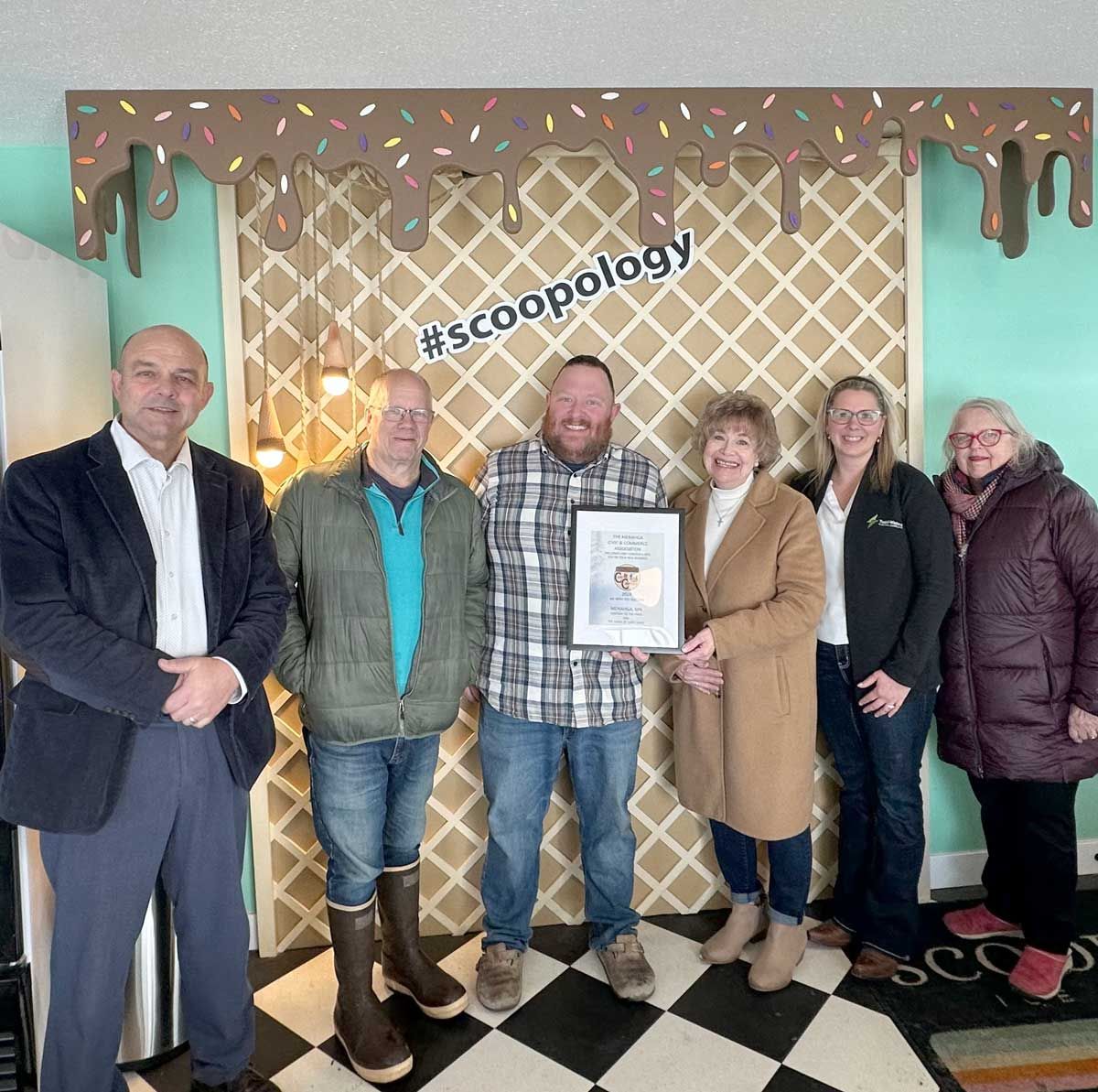 Six people stand in front of a waffle-patterned wall under a chocolate-drip decoration, one holding a framed certificate.