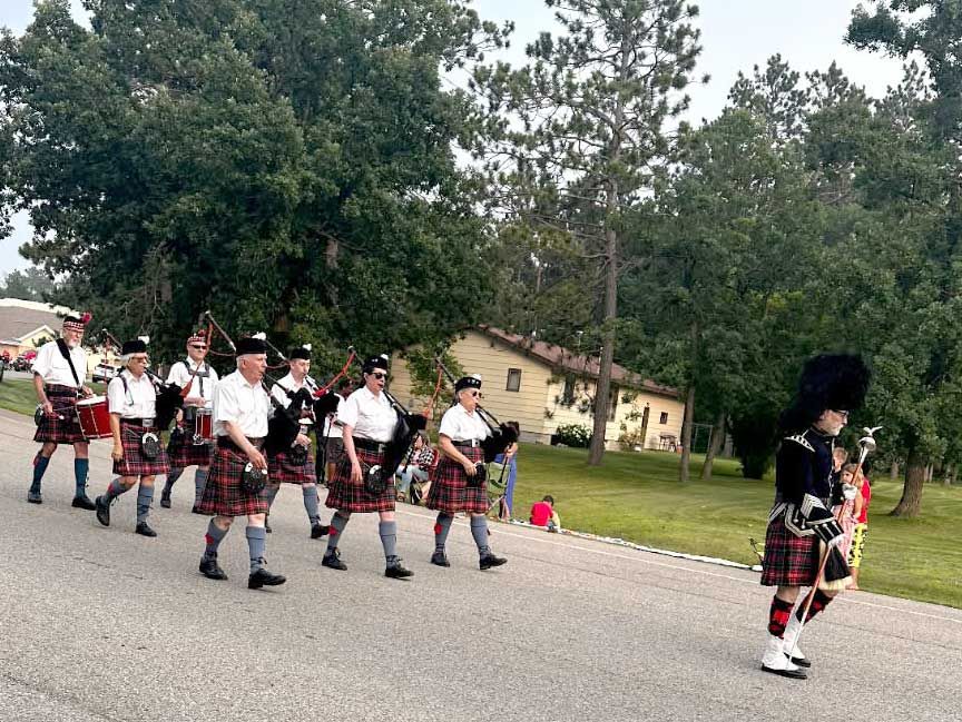 A line of bagpipers in kilts marching on an asphalt road in front of trees and a building.