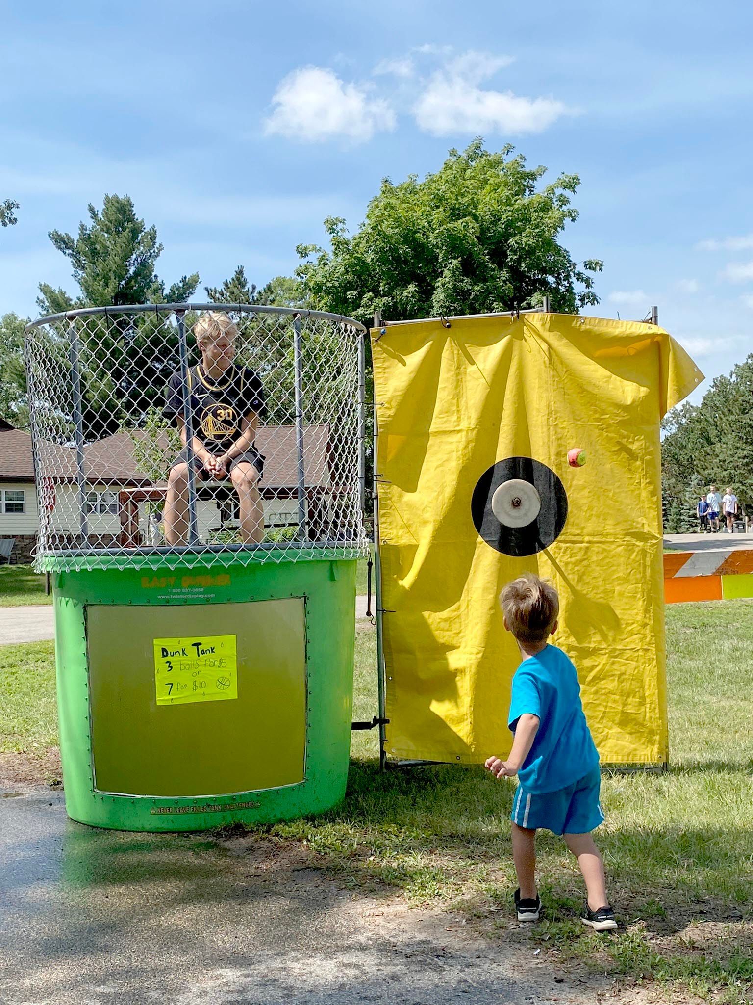 A person sits in a dunk tank while a child prepares to throw a ball at the target during an outdoor event.