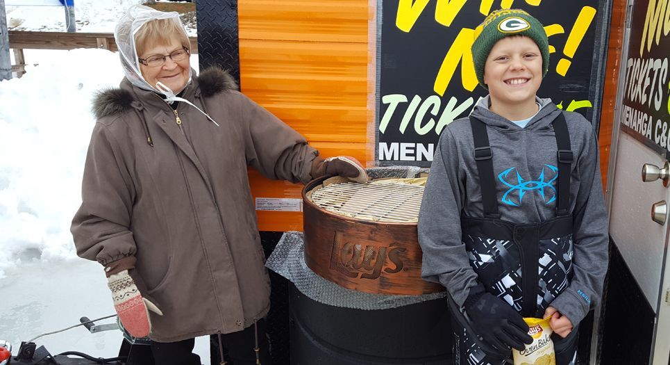 An older adult and a youth stand outdoors in snowy weather by a ticket booth next to a large circular container of minnows.