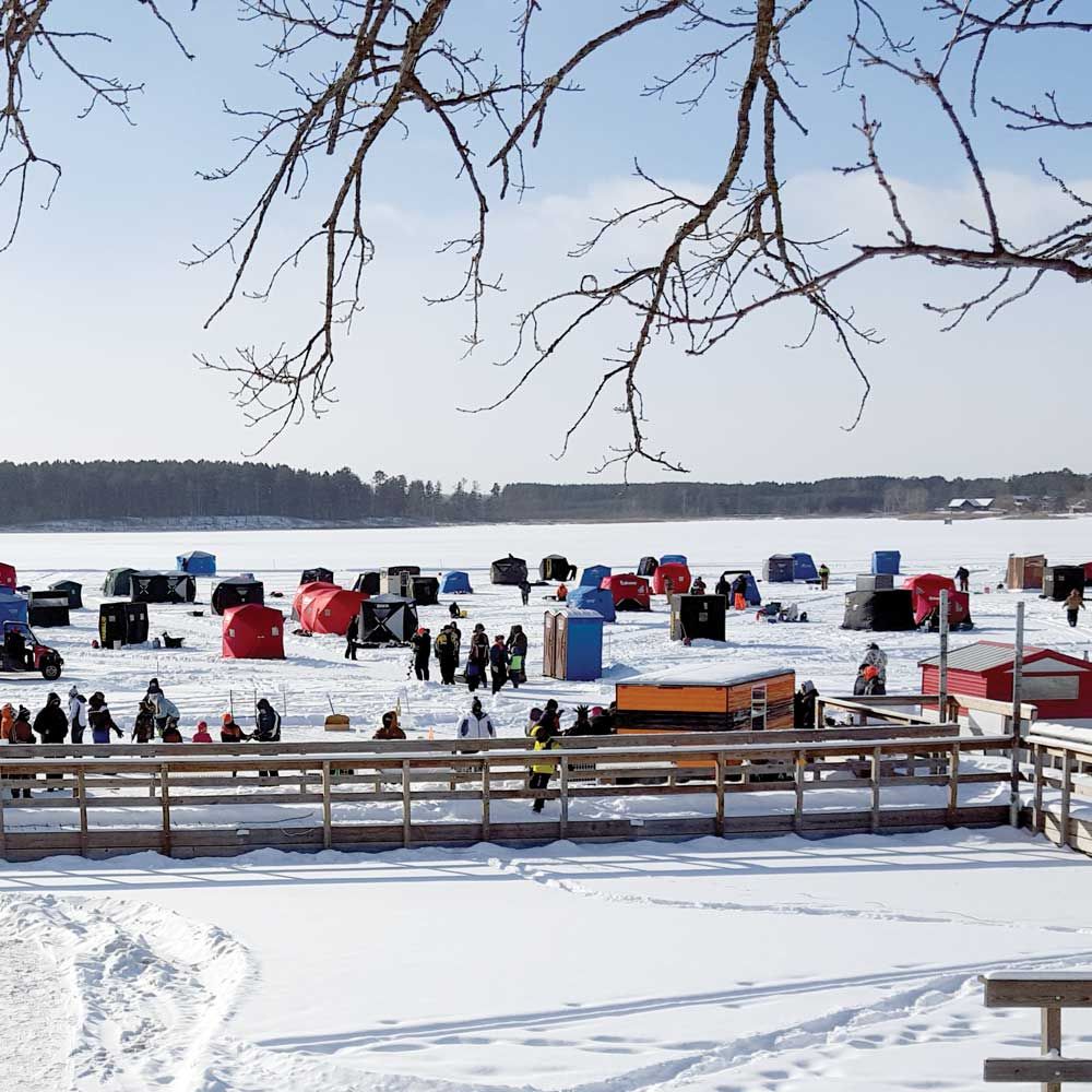 A frozen lake filled with numerous colorful ice fishing shelters and people on a sunny winter day, viewed from a deck.