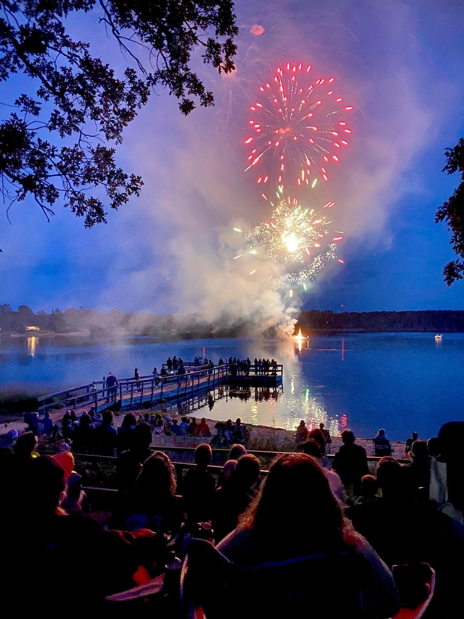 Crowd gathered on a lakeside dock at twilight watching colorful fireworks explode in the sky.