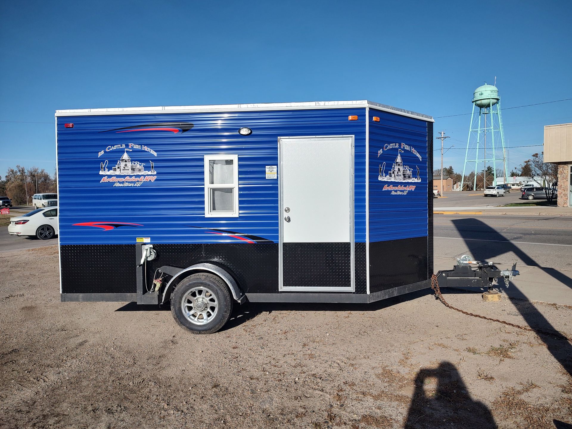 A blue and black enclosed cargo trailer parked on a gravel lot with a water tower in the background.
