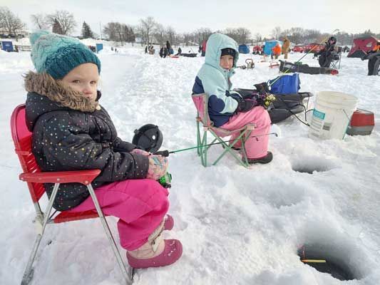 Two children in winter gear sit on small folding chairs on a frozen, snow-covered lake, ice fishing near open holes.