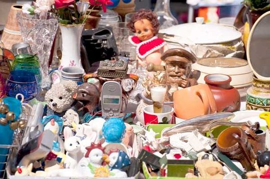 A cluttered table at a flea market filled with diverse vintage items, including glassware, dolls, ceramics, and ornaments.