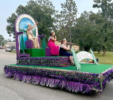 A parade float decorated in purple and green with three individuals wearing crowns and sashes riding on tiered platforms.
