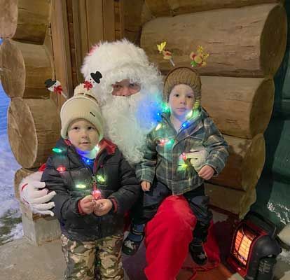Santa Claus sits with two children in a log cabin, all wearing holiday-themed hats and festive string lights.