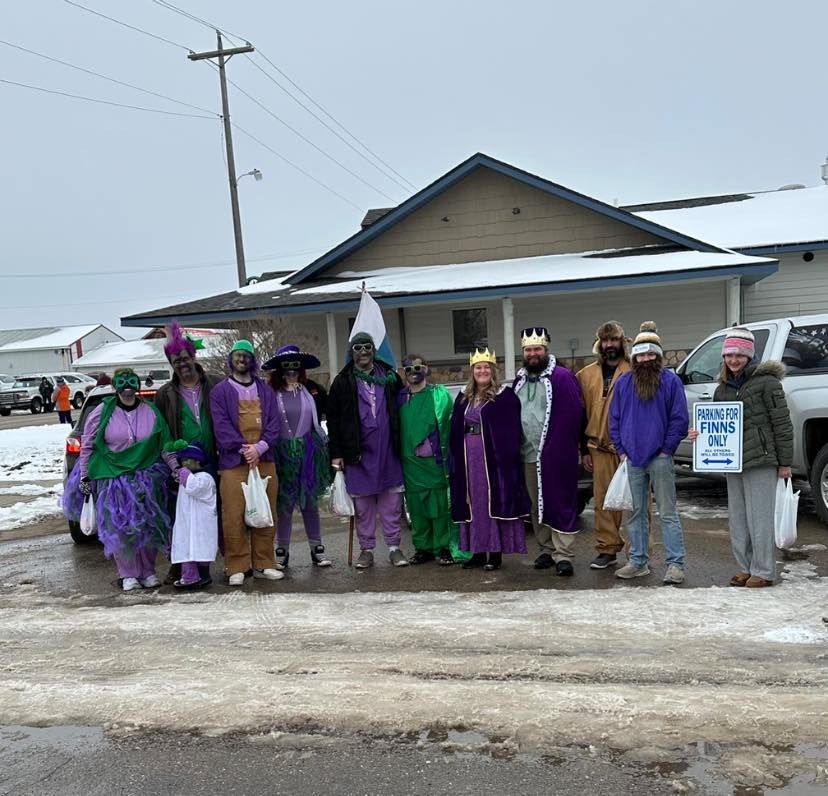 A group in purple and green costumes stands outside a snowy building, one holding a sign.