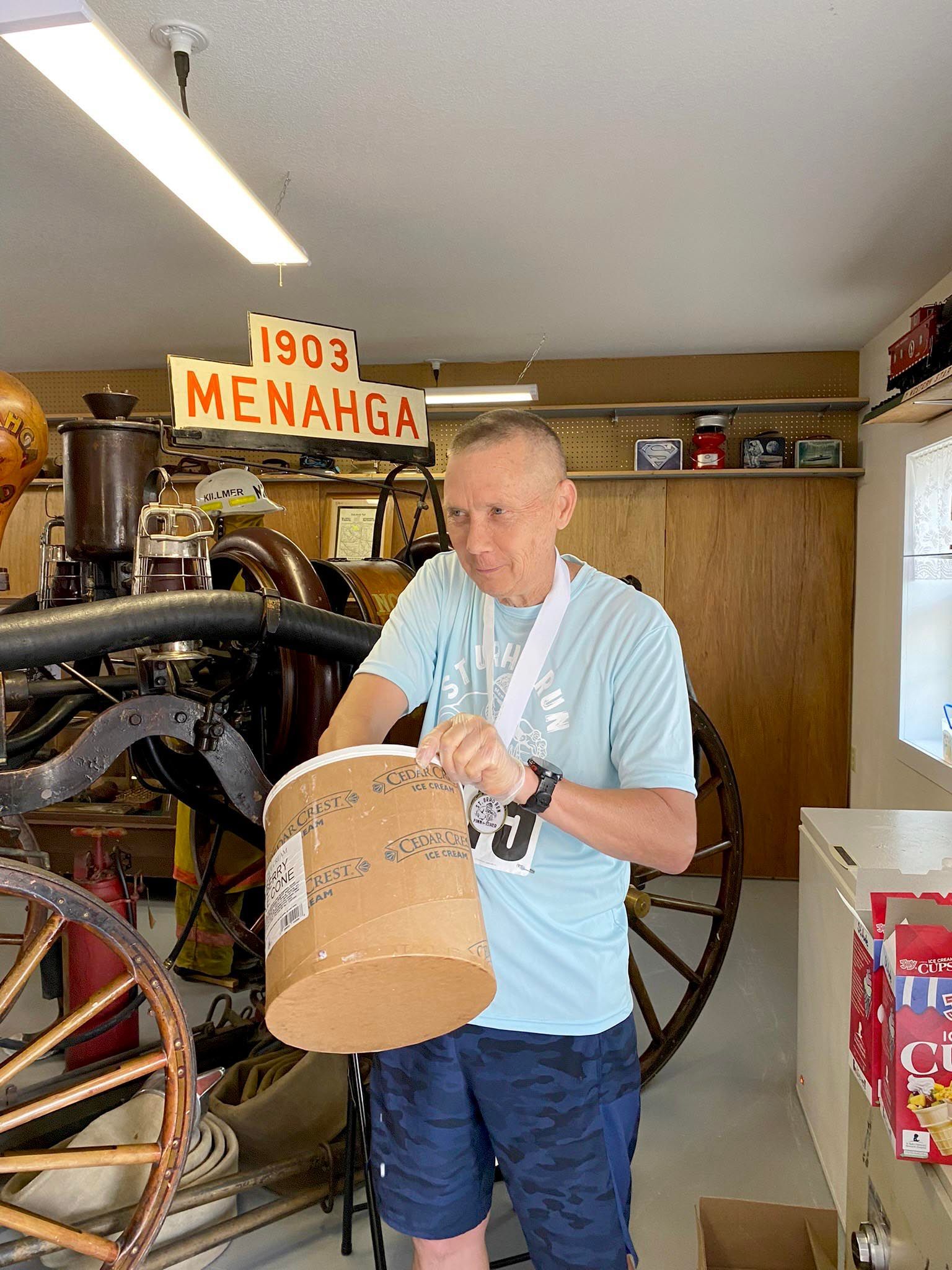 A person in a light blue shirt holds a round cardboard box in front of a 1903 Menahga fire engine display.