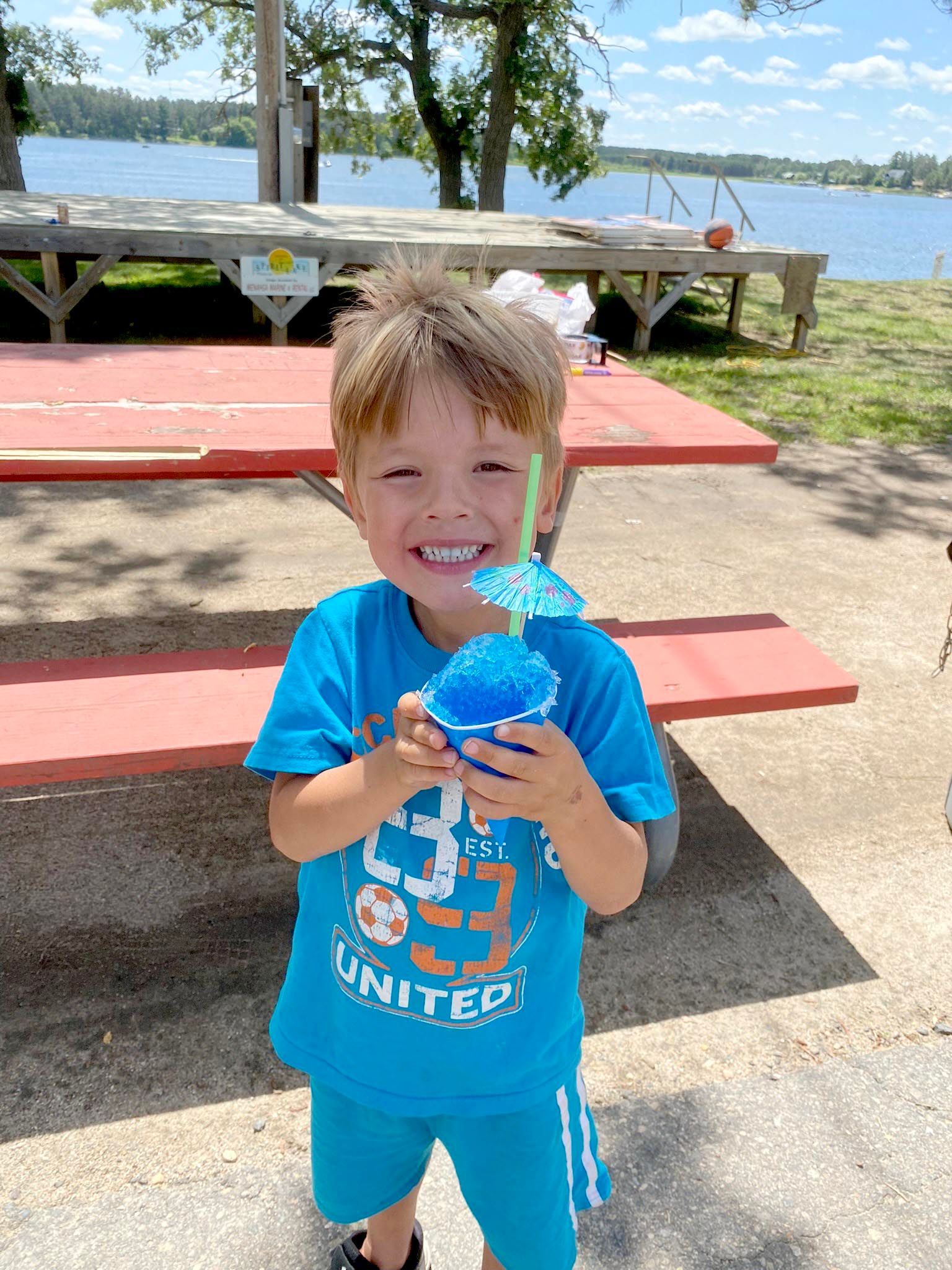 A smiling person in a blue shirt holds a blue shaved ice with a small paper umbrella at a lakeside picnic table.