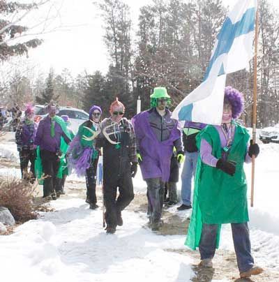 A group of people wearing costumes, masks, and colorful wigs walk through snow, one carrying a Finnish flag.
