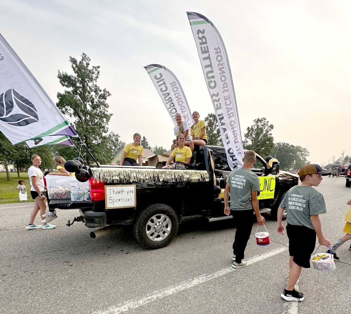 A black truck decorated for a parade carries people and sponsor banners in a parking lot, with attendees nearby.