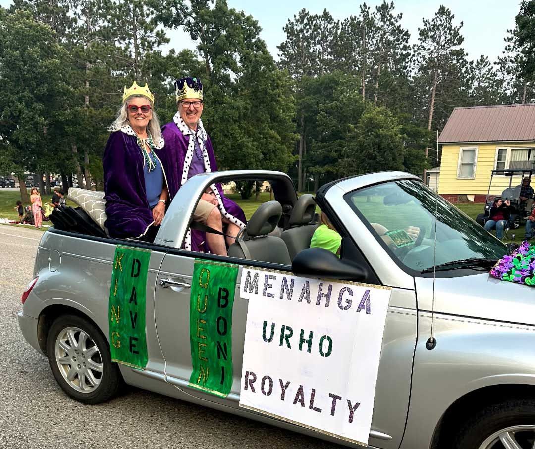 Two people in crowns and purple capes ride in a silver convertible marked 