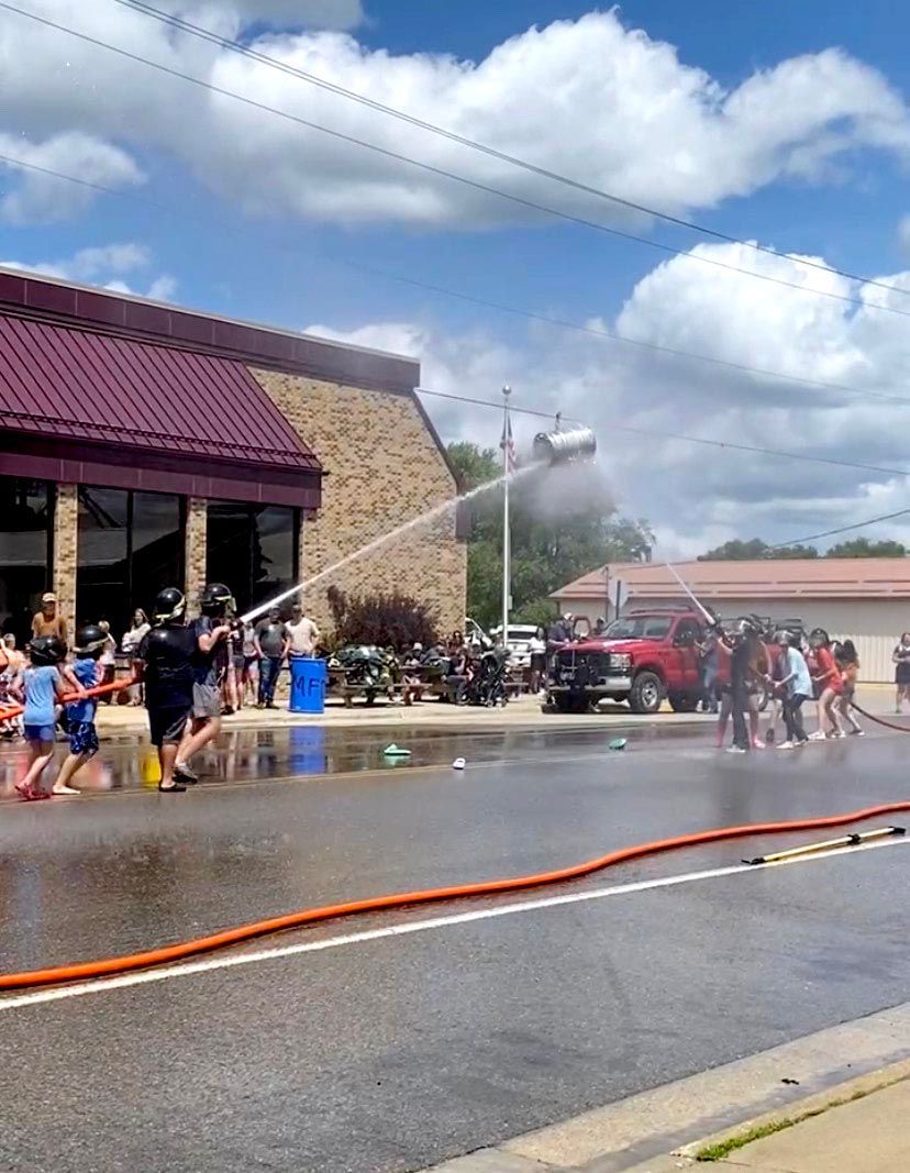 People participate in a fire hose challenge on a sunny street near a brick building, spraying water at a suspended barrel.