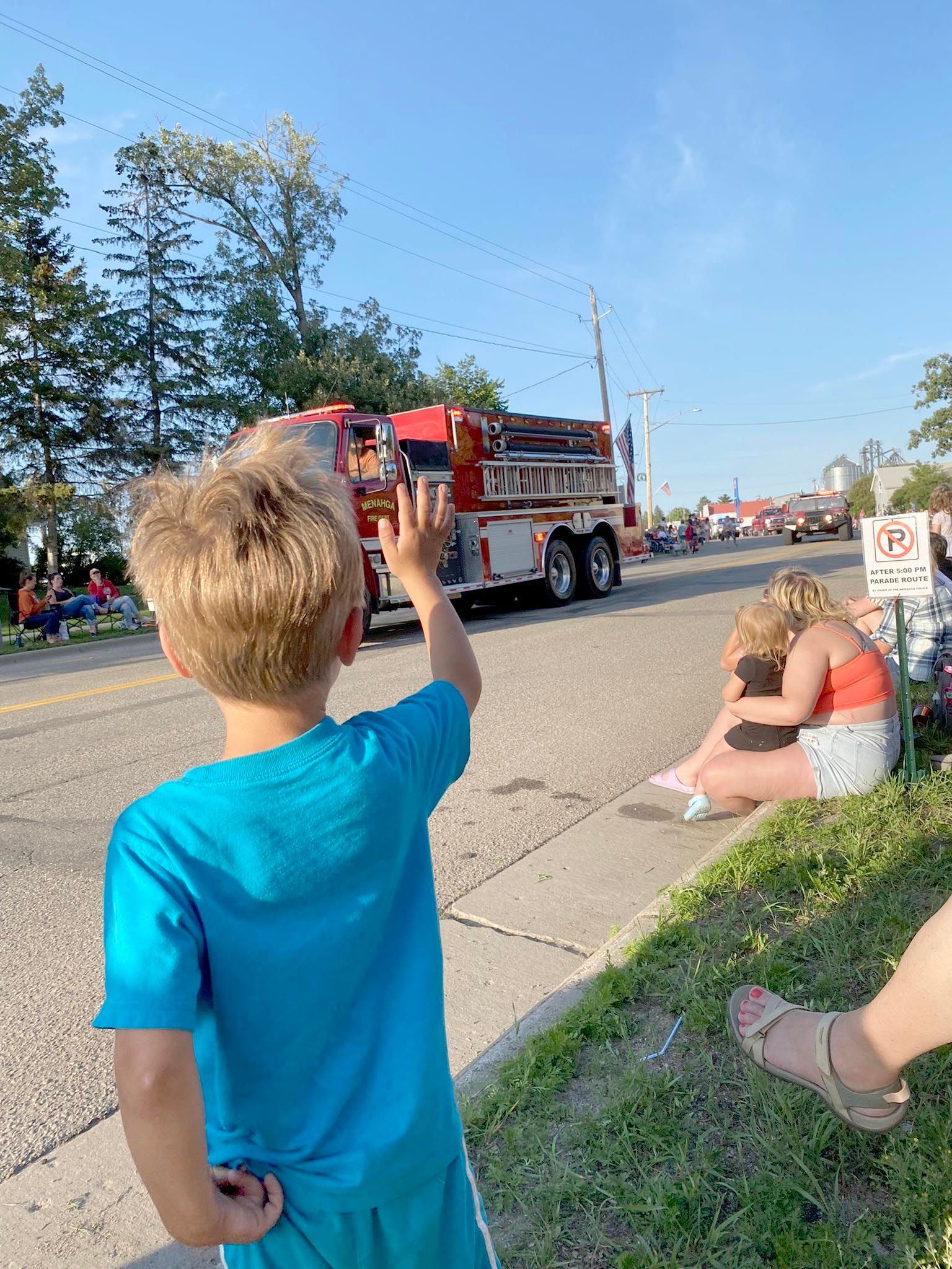 A child in a blue shirt waves at a passing fire truck during an outdoor parade, with people watching from the grass.