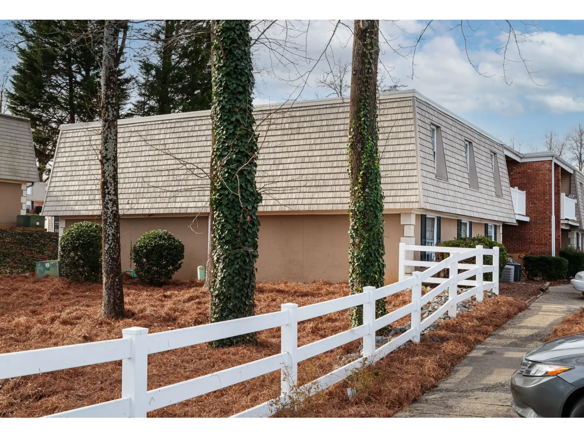 Exterior view of a beige apartment building with a white fence, ivy-wrapped trees, and trimmed bushes.