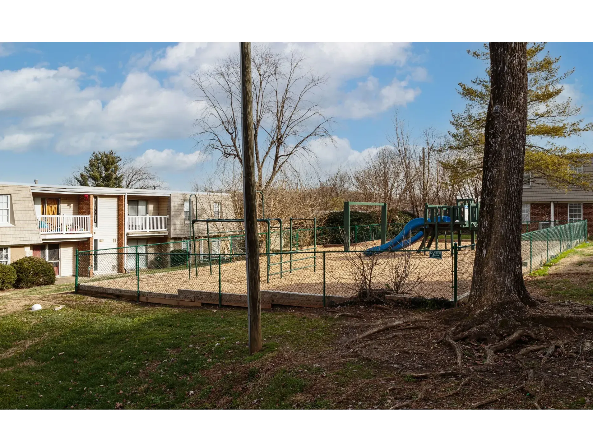 Outdoor playground area at an apartment community with a blue slide, fencing, and nearby buildings.
