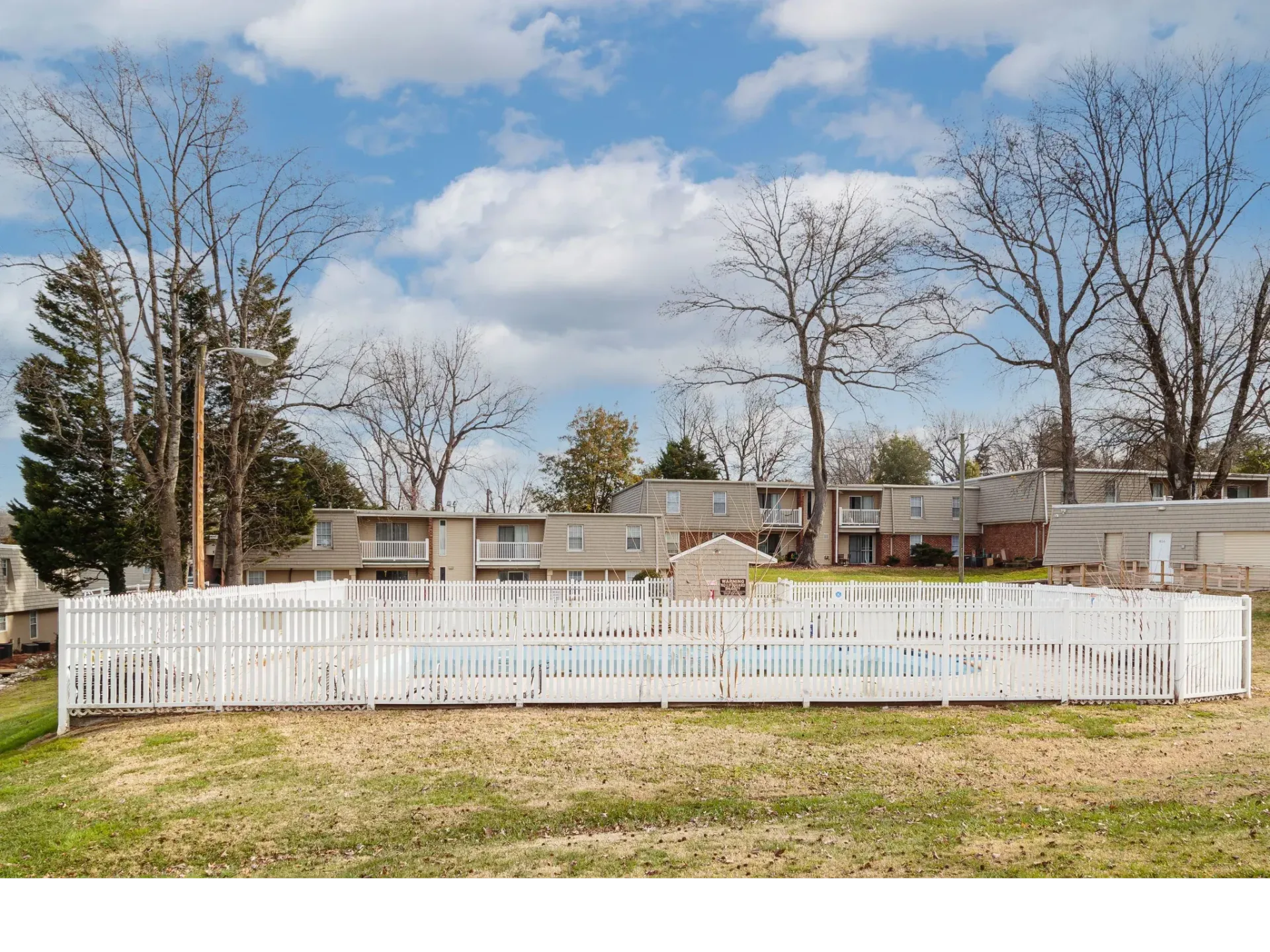 Fenced community pool with low-rise apartment buildings and bare trees under a blue sky.