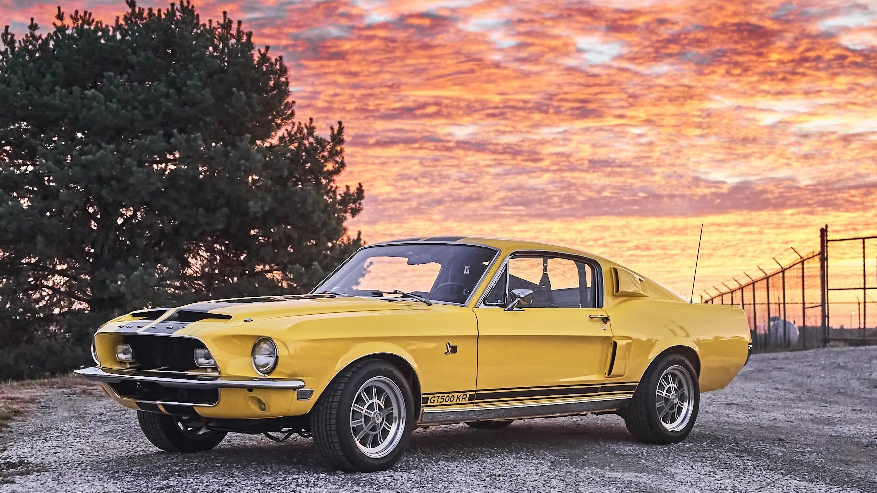 A yellow mustang is parked in a gravel lot at sunset.
