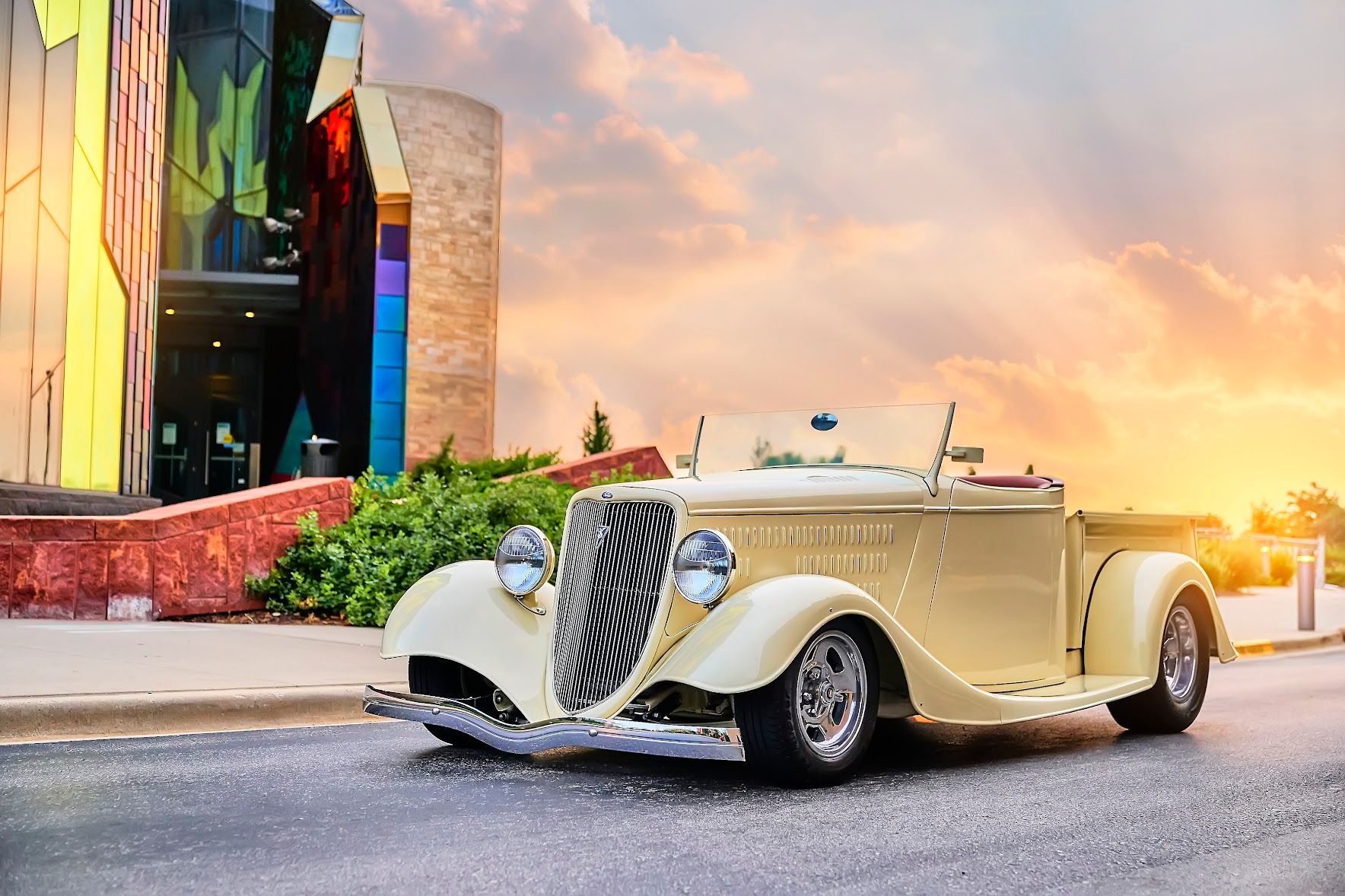A vintage car is parked on the side of the road in front of a building at sunset.