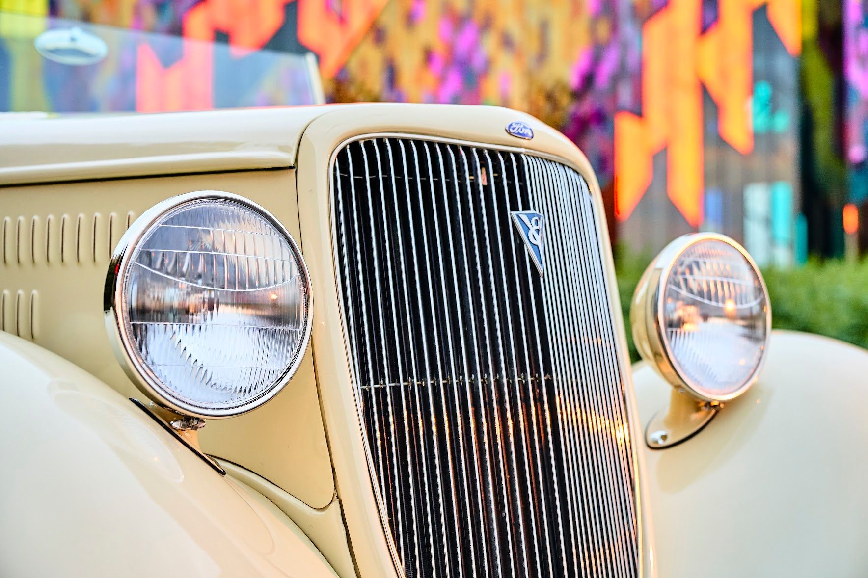 A close up of the front of an old car with a graffiti wall in the background.