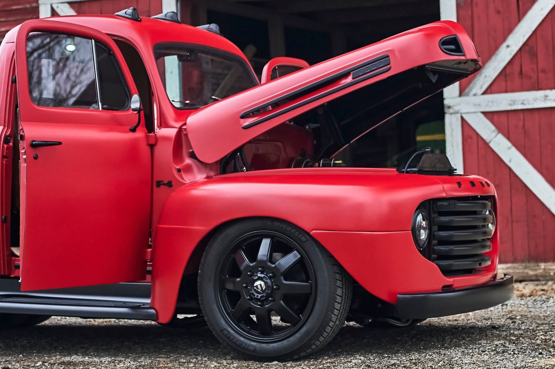 A red truck with the hood open is parked in front of a barn